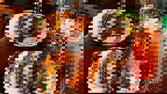 Two plates of roast dinner with meat, potatoes, Yorkshire pudding, and vegetables on a wooden table by a fireplace, accompanied by red wine, Sol beer, and a cocktail with a lemon garnish.