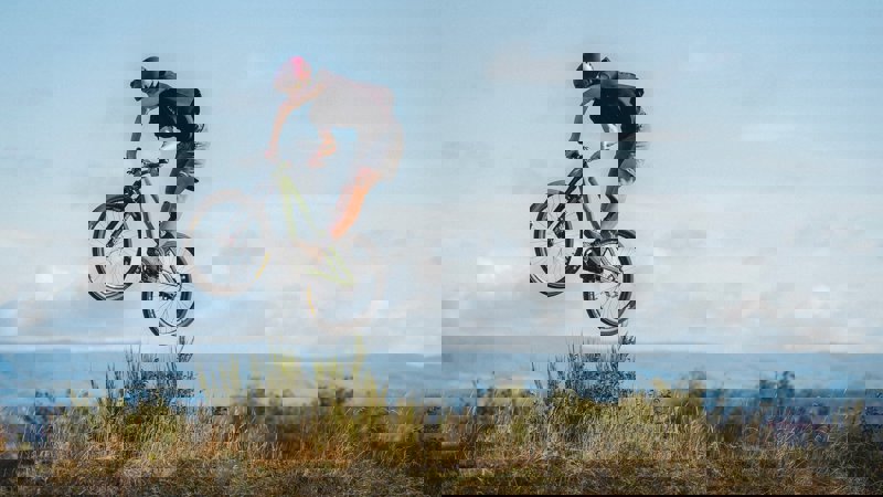 A cyclist mid-air performing a jump on the mountain bike trails at Cathkin Braes, with hills and sky in the background.