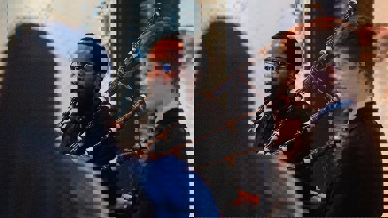 Two men are playing bagpipes indoors. One man has a beard and glasses, and the other is smiling.