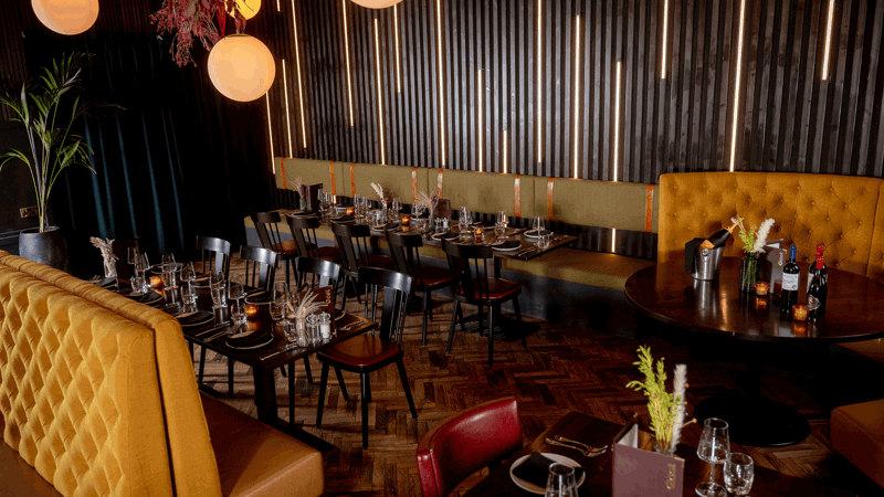 Modern restaurant interior with mustard yellow booths, black chairs, wooden tables, pendant lights, vertical slat walls, and table settings including glassware, plates, and cutlery.