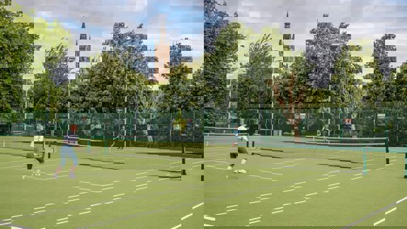 An outdoor tennis court with two people playing, and a large building with a tower in the background.