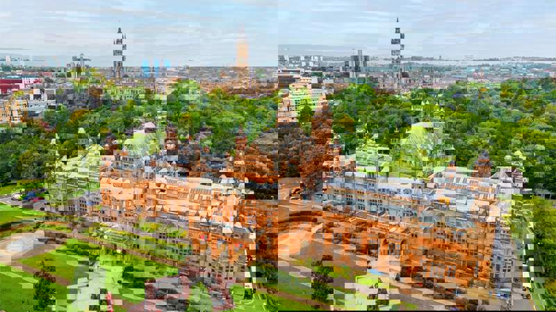 Drone view of a grand red building surrounded in greenspace, with a grand building with a spire and hills in the distance.