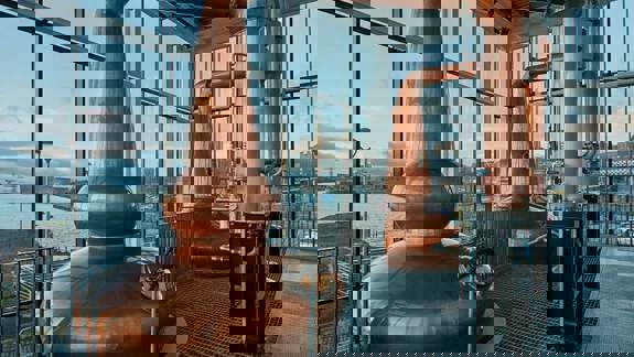 A modern distillery with two large, shiny copper stills and large windows looking out onto water.