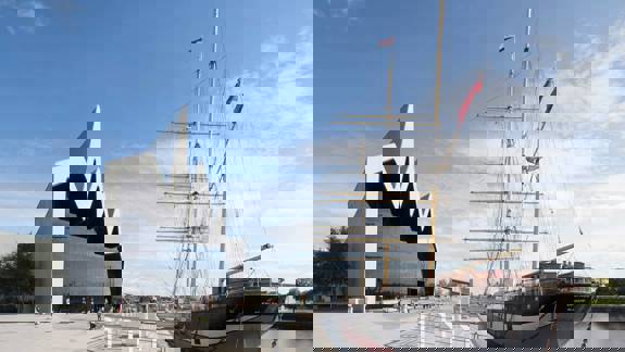 A modern, angular building with a jagged roofline sits next to a historic, three-masted ship docked on a river.