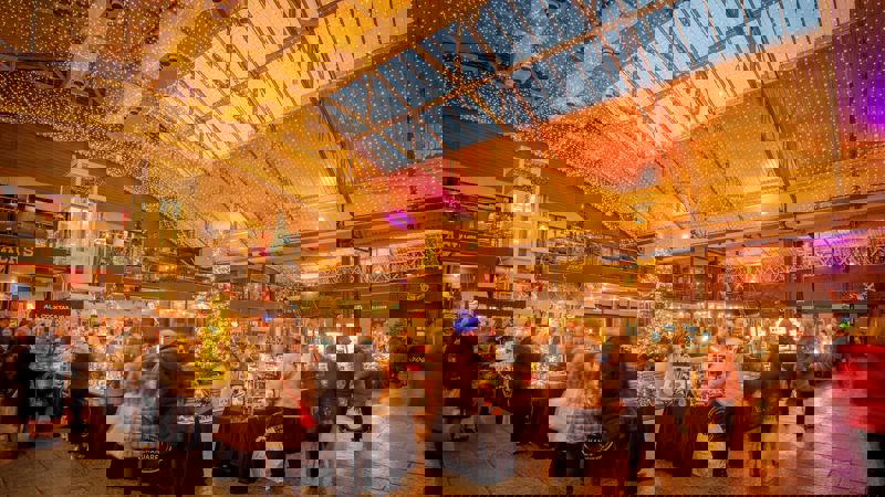 A wide shot of an indoor market with a vaulted glass ceiling, decorated with many small lights and a Christmas tree.