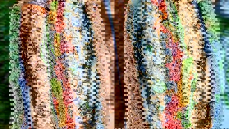 A close-up of a thick sandwich, sliced in half, revealing a creamy egg salad filling with avocado, tomato, and lettuce, on wholemeal bread.