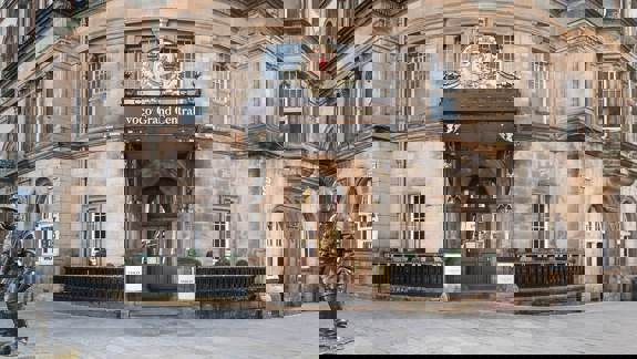 A historic, stone-built hotel in Glasgow, showing a central ornate doorway with a royal crest and a statue of a helmeted figure.