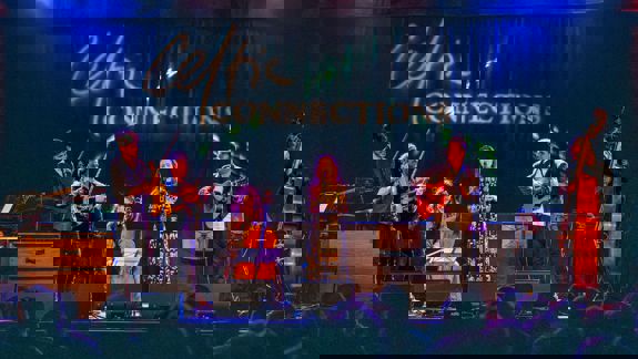 A band performs on a stage with various instruments, including string instruments and guitars, with "Celtic Connections" projected on the stage backdrop.