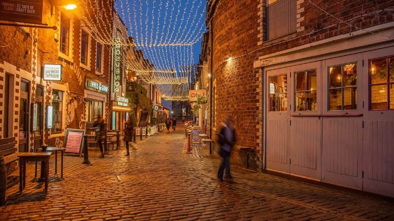 A cobblestone lane in Glasgow, Ashton Lane, lit by hanging fairy lights and lanterns, with people walking along it at dusk.
