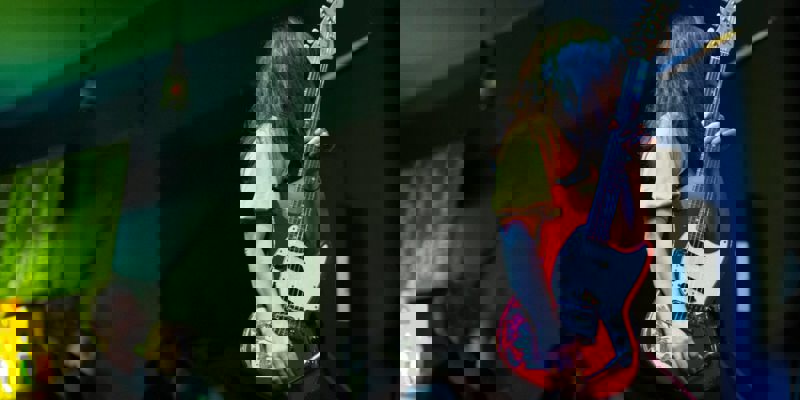 A musician with long dark hair plays a red electric guitar on a dimly lit stage, with blurred lights in the background.