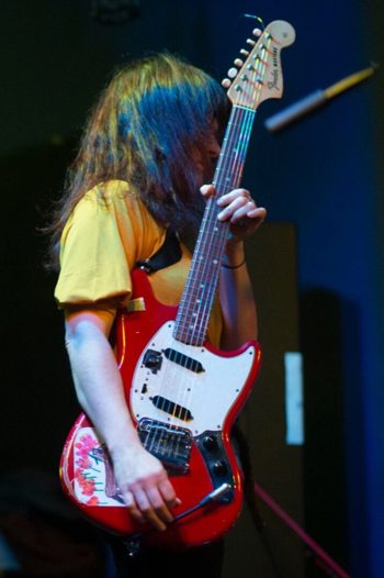 A musician with long dark hair plays a red electric guitar on a dimly lit stage, with blurred lights in the background.