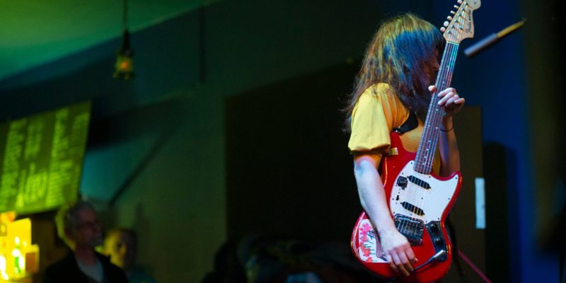 A musician with long dark hair plays a red electric guitar on a dimly lit stage, with blurred lights in the background.