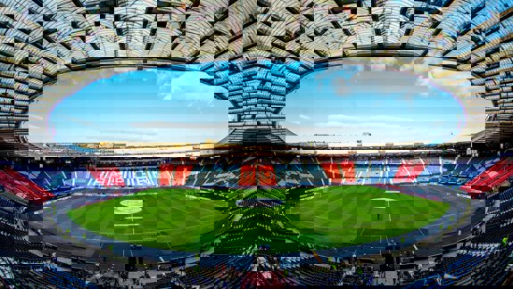 An empty Hampden Park stadium with blue and red seats, a green field, and a transparent roof against a blue sky.