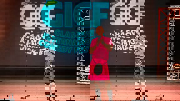 A smiling comedian in a pink shirt and kilt is on stage at the Glasgow International Comedy Festival (GICF) with a microphone.