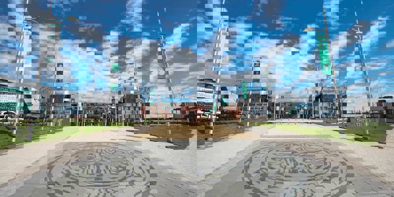 An wide view of the entrance to Celtic Park stadium, with a large Celtic FC crest on the ground and banners.