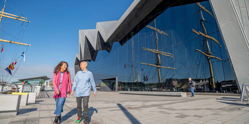 Two people walk and talk outside a modern museum with a striking zigzag roof and glass walls reflecting a tall ship under a clear blue sky.