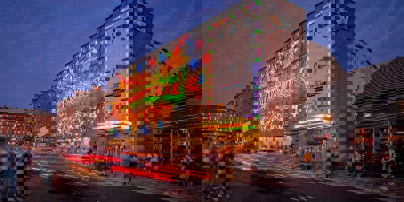 A street-level view of the Barrowland Ballroom at night. The building is lit with a colourful neon sign with stars and the name "Barrowland" in a large font. The long exposure shot shows blurred light trails from cars on the street.