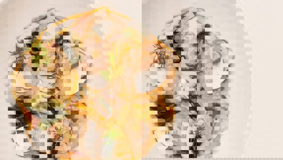 A close-up overhead shot of a white bowl filled with thinly sliced scallops, mushrooms, seaweed, and garlic in a savoury sauce, garnished with fresh dill.