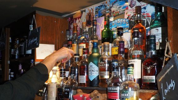 A hand reaching for a whisky bottle on a bar shelf filled with various Scotch whisky brands, labeled by region.