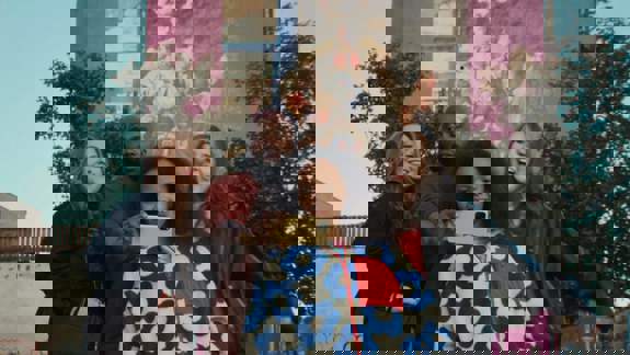 A group of four smiling friends take a selfie together outdoors. They stand before a pink building featuring a large mural, surrounded by green trees under a clear sky.