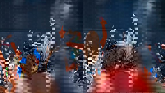 Concert crowd with people raising their arms and phones toward the stage, viewed from behind two women sitting on shoulders.