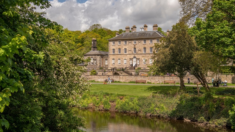 Pollok House is captured from across the a river. Trees are on either side and the sun is shining. 