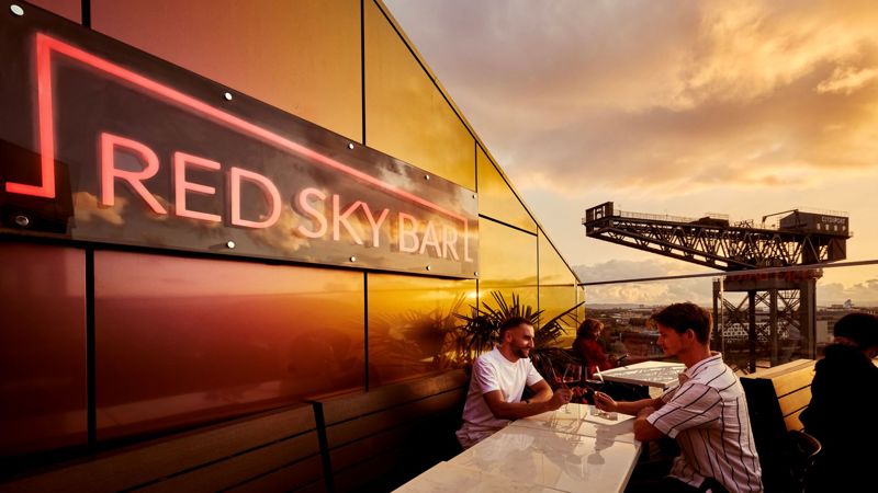 Outdoor seating area at Red Sky Bar with a neon sign on the wall, people sitting at a table, and a view of a large industrial crane against a sunset sky