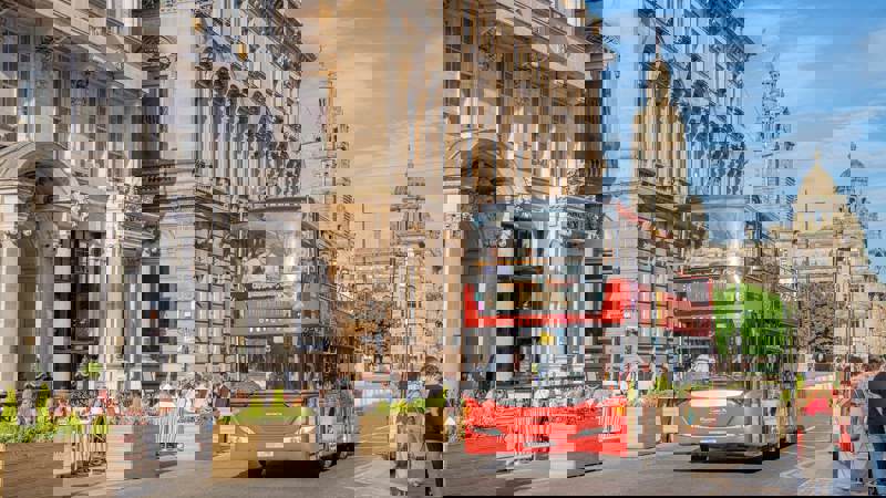 A red double-decker sightseeing bus drives through a sunny street in Glasgow with historic buildings and people dining outdoors.