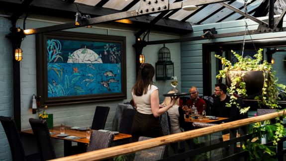 A waiter serves a couple seated in the bright, modern conservatory area of the Ubiquitous Chip, with a large painting on the wall.