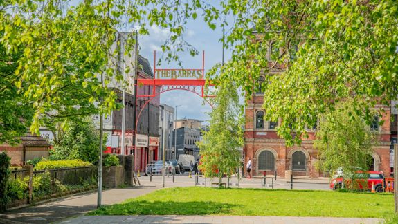 A sunny, green view frames a red archway sign reading "THE BARRAS" in the distance, with a Victorian brick building on the right.