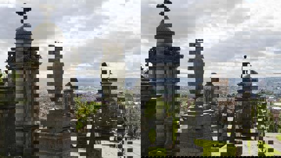 An old cemetery filled with elaborate stone monuments and gravestones, overlooking a city skyline under a cloudy sky.