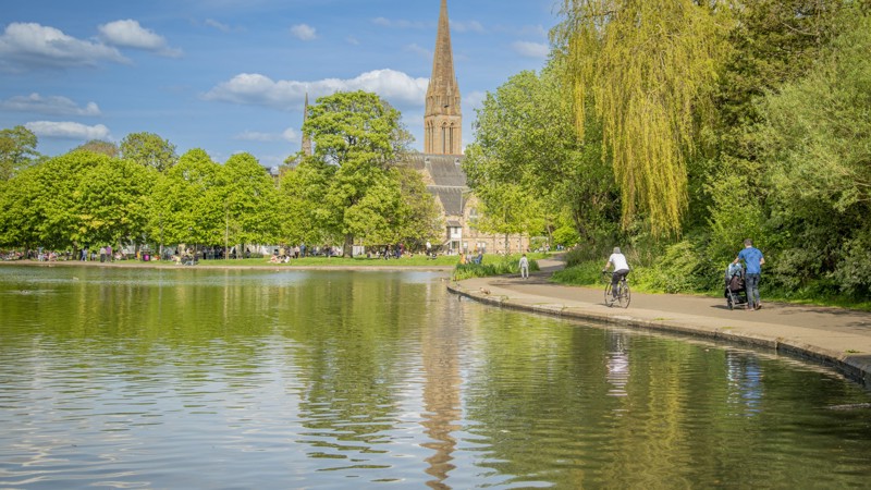 People walk and cycle around a pond in a city park on a sunny day. Trees and a church spire surround the pond.