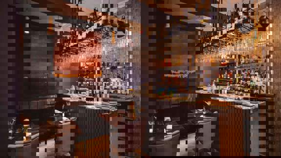 A dimly lit restaurant bar with a seating area. There is an abstract painting on the wall and a fully stocked bar with hanging wine glasses.