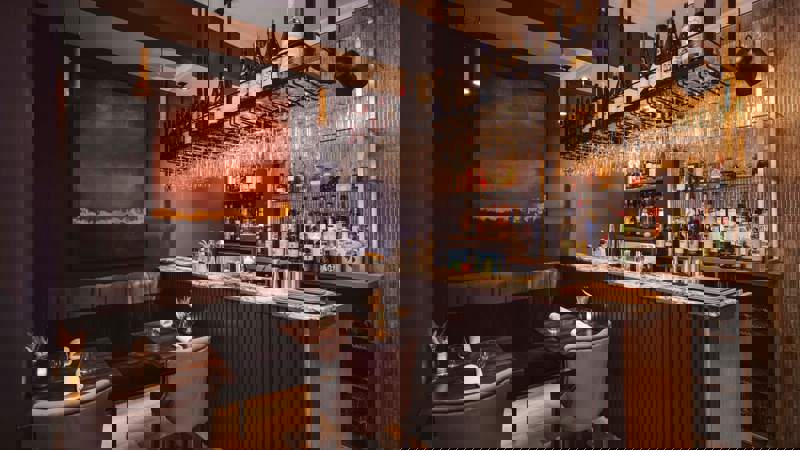 A dimly lit restaurant bar with a seating area. There is an abstract painting on the wall and a fully stocked bar with hanging wine glasses.