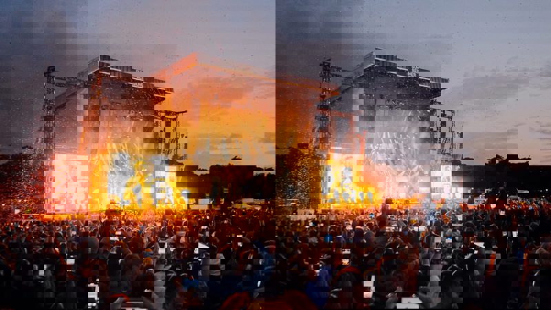 A night time crowd holding up phones to capture a performance on a large, brightly-lit stage with pyrotechnics.