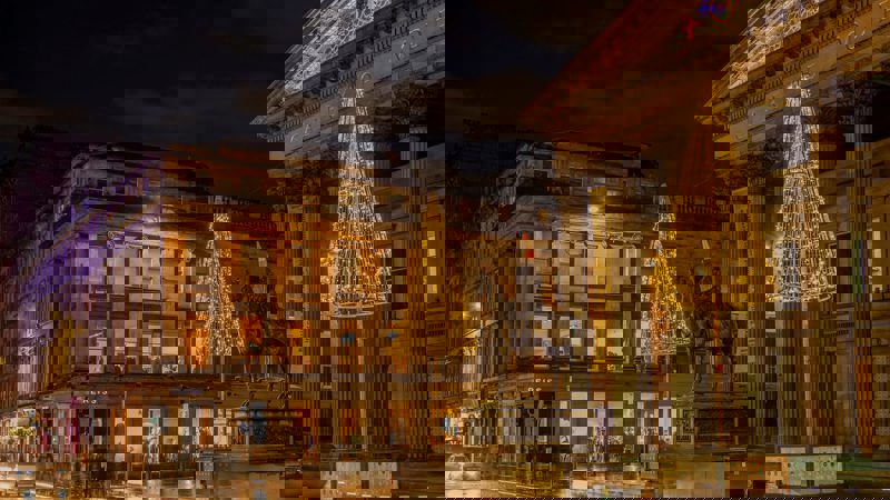 A festive nighttime view of the Gallery of Modern Art and the Duke of Wellington statue in Glasgow, adorned with golden Christmas lights and glowing trees.