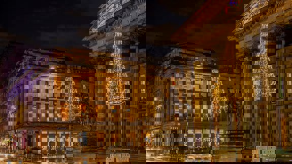 A festive nighttime view of the Gallery of Modern Art and the Duke of Wellington statue in Glasgow, adorned with golden Christmas lights and glowing trees.