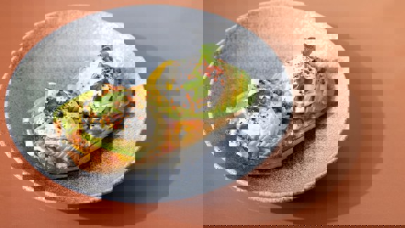 Close-up of a brunch dish of avocado toast with poached eggs and chilli flakes on a grey plate against a peach coloured table.