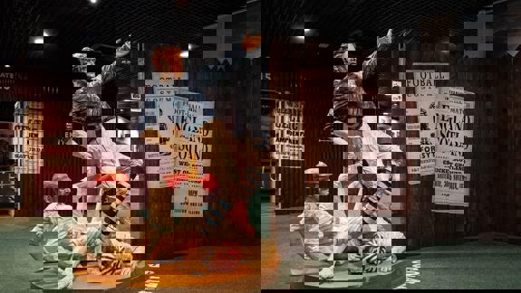 A life-size statue depicts two 19th-century footballers tackling each other, with one player falling and the other standing over him. In the background are historical posters and displays.