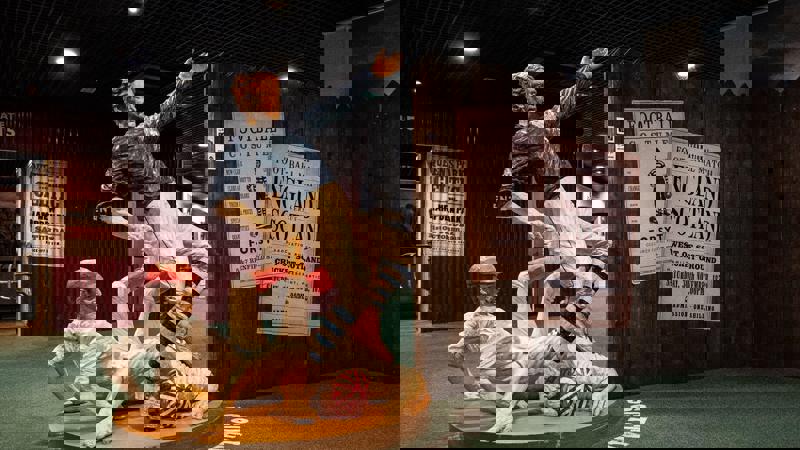 A life-size statue depicts two 19th-century footballers tackling each other, with one player falling and the other standing over him. In the background are historical posters and displays.