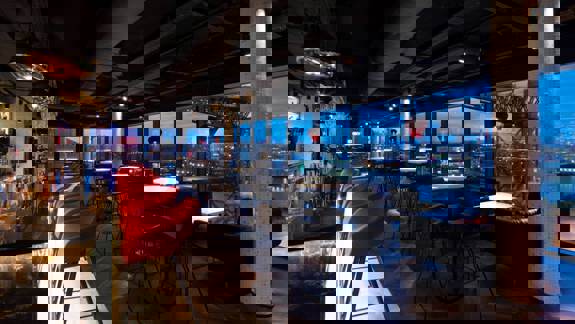 A rooftop bar with floor-to-ceiling windows showcasing city skyline views, modern furnishings in dark tones, and a lively crowd gathered around sleek tables.