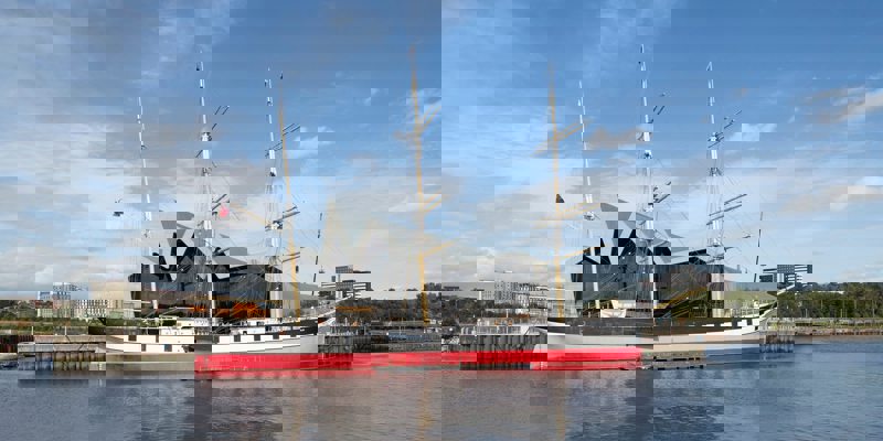  The Glenlee tall ship is docked on the River Clyde in front of the modern, zigzag-roofed Riverside Museum in Glasgow under a bright blue sky.