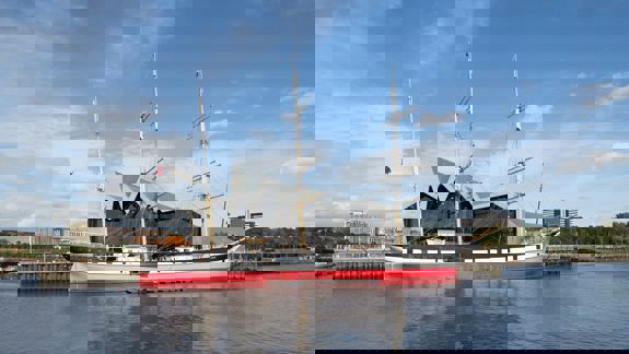 The Glenlee tall ship is docked on the River Clyde in front of the modern, zigzag-roofed Riverside Museum in Glasgow under a bright blue sky.