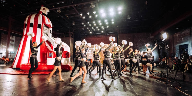 Cheerleading group with pom-poms performs indoors in formation beside a large red and white inflatable structure under bright stage lights.