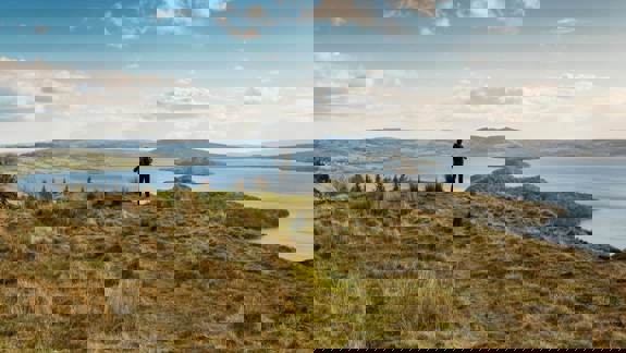 A hiker stands on a grassy hill overlooking a vast loch dotted with islands, with rolling hills in the background
