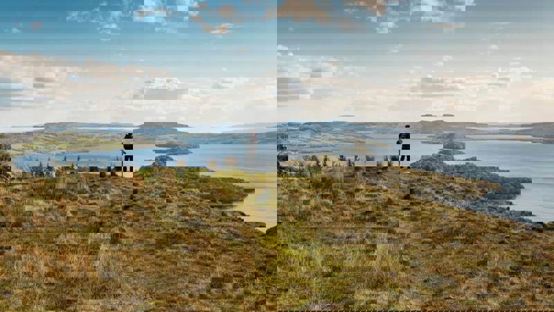 A hiker stands on a grassy hill overlooking a vast loch dotted with islands, with rolling hills in the background