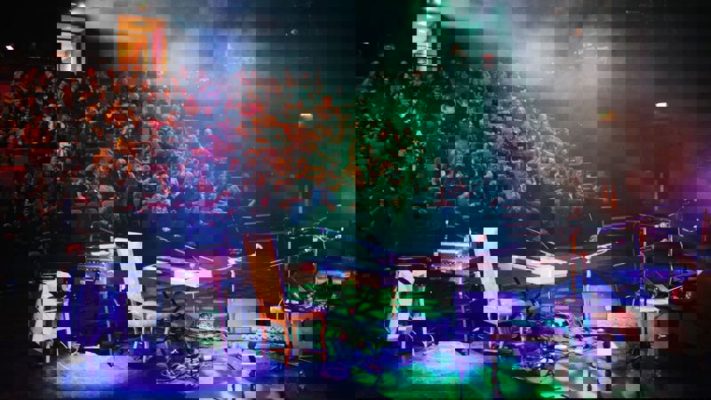 An audience seated in a dark theatre watches a performance on a smoke-filled stage lit with colourful lights, with musical instruments in the foreground.