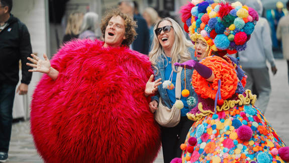 A laughing festival attendee stands with costumed performers, one dressed in a big, shaggy pink ball suit.