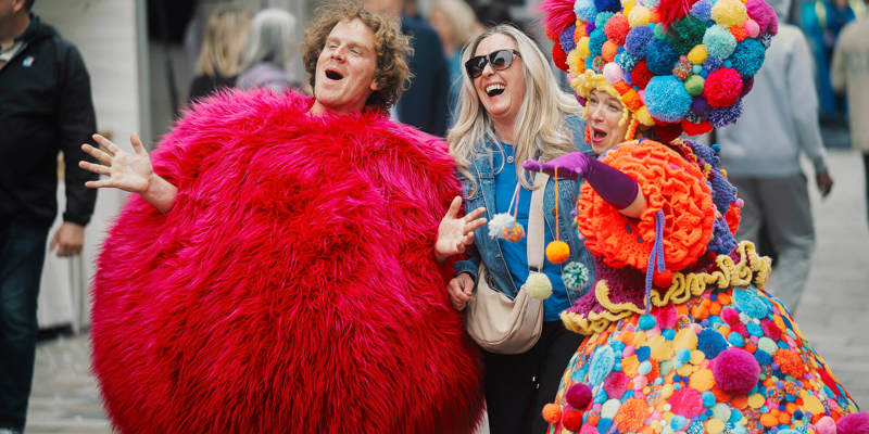 A laughing festival attendee stands with costumed performers, one dressed in a big, shaggy pink ball suit.
