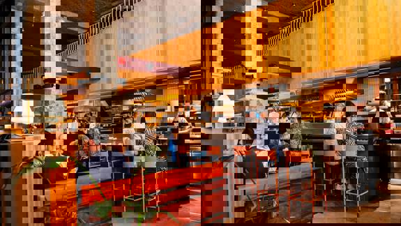 The interior of a modern restaurant with a counter, people seated at tables and a chef working in the open kitchen.